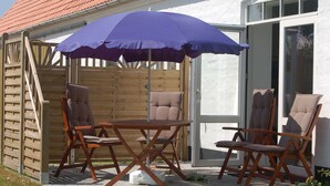 Een terras met een houten tafel en vier stoelen, een blauwe parasol en een houten omheining.