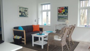 Een moderne woonkamer met een donkergrijze bank, een witte salontafel, rieten stoelen en een kleine eettafel. Twee ramen zorgen voor natuurlijk licht, en een kleurrijk schilderij hangt aan de muur.