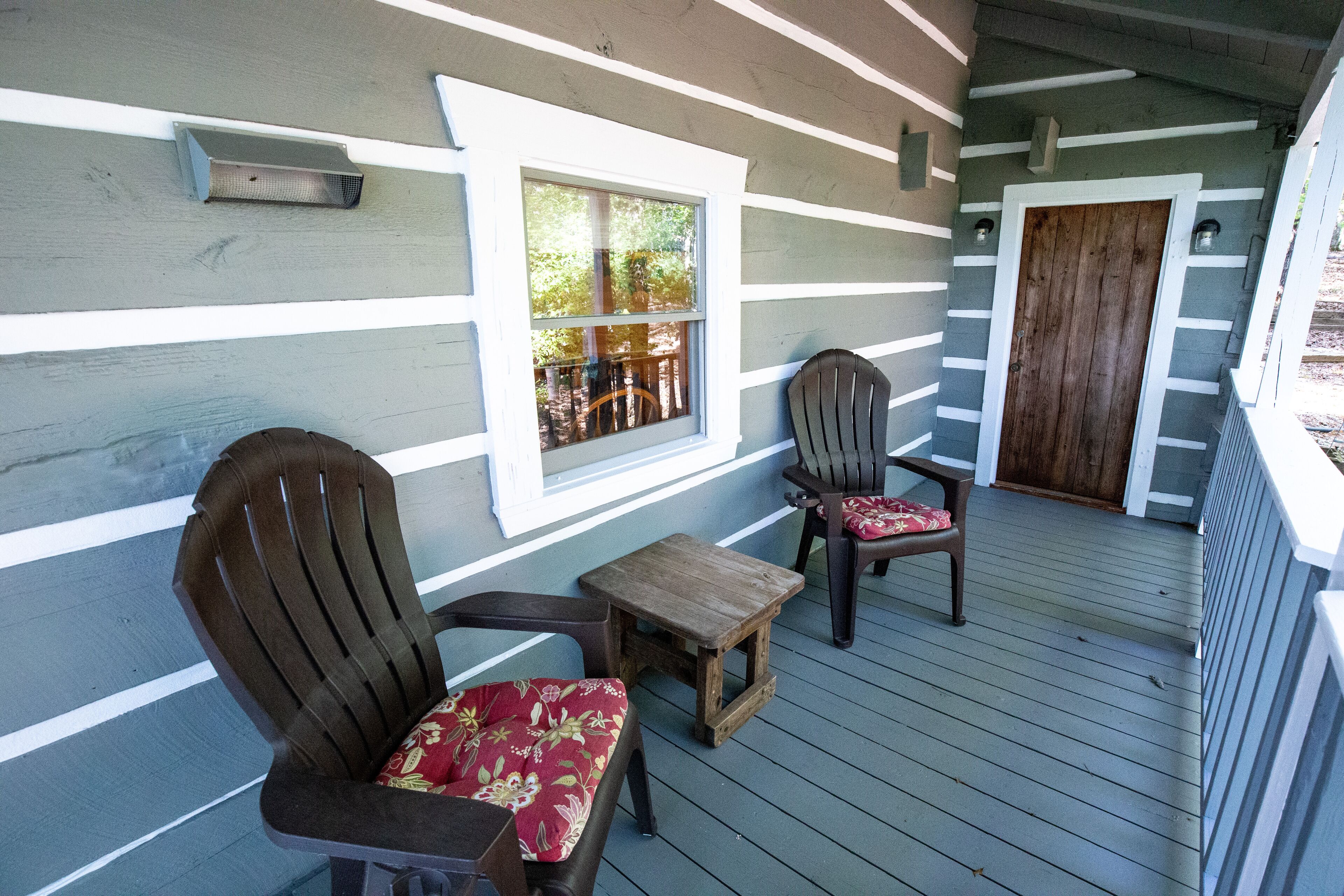 A wooden deck with two wooden chairs and a small table, a window with a view of trees, and a wooden door.