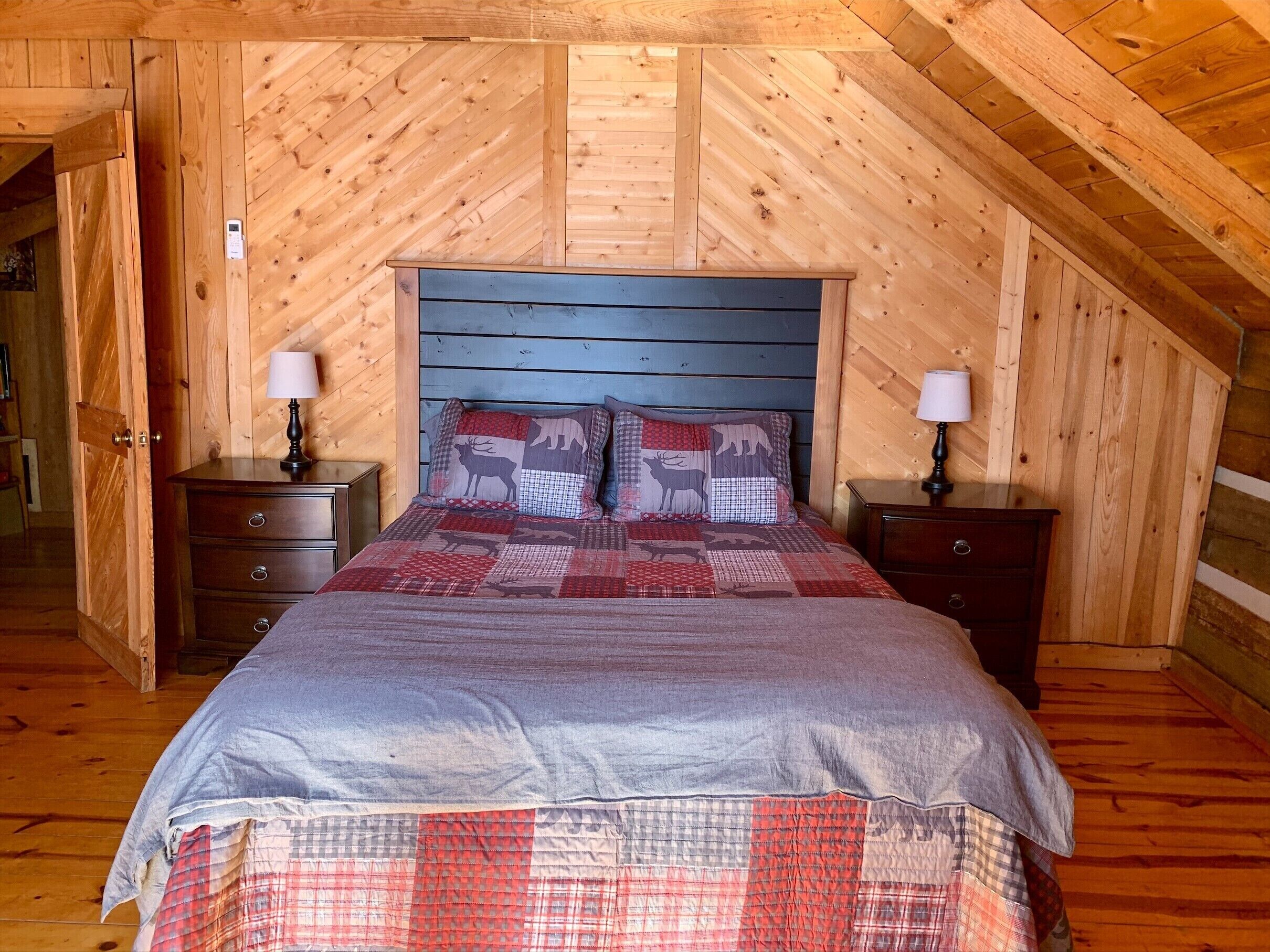 Een gezellig slaapkamer onder een schuin houten dak, een bed met een geruit dekbed, twee nachtkastjes met lampen, en een houten garderobe.
