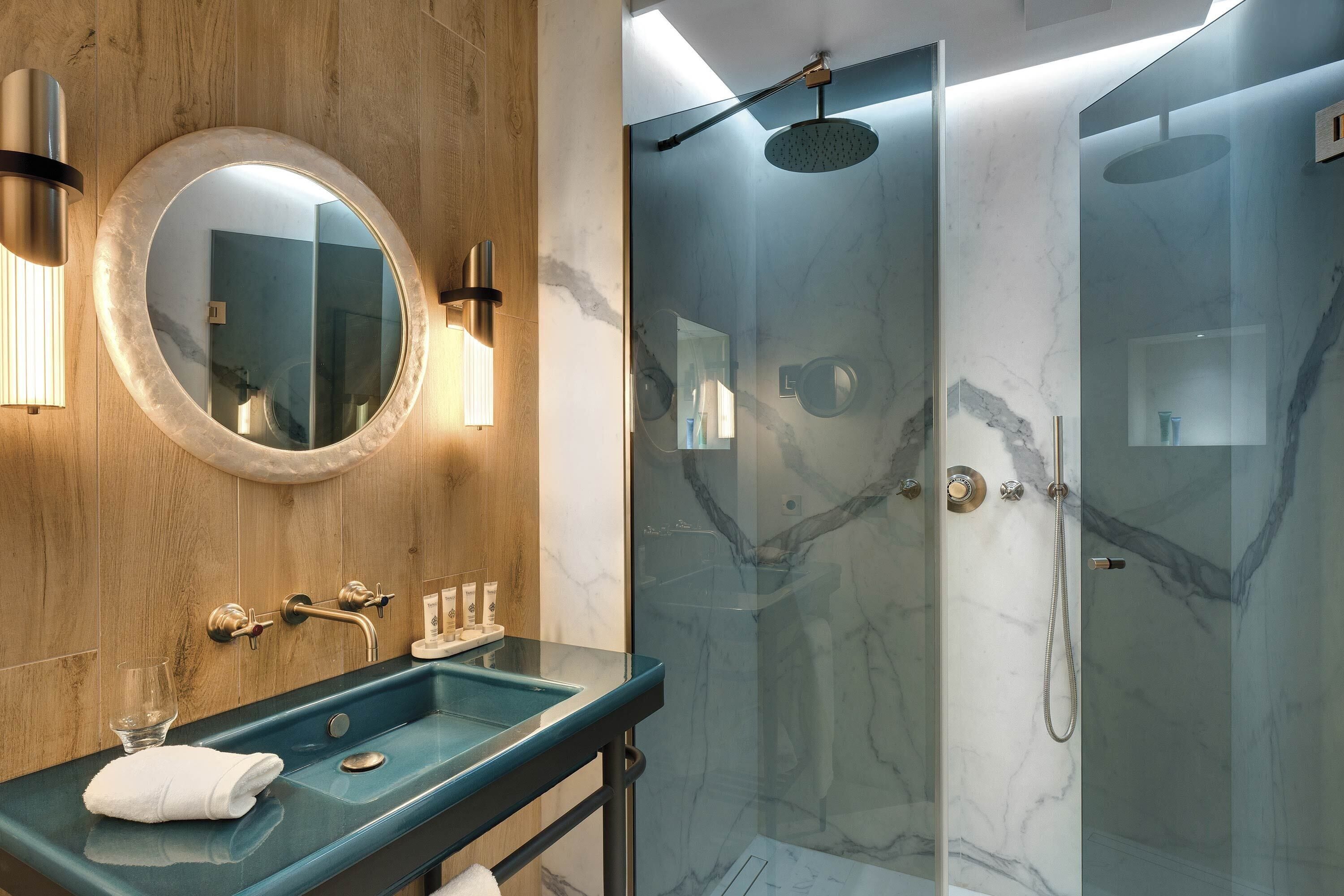 A modern bathroom with a marble sink, a round mirror, and a glass-enclosed shower.