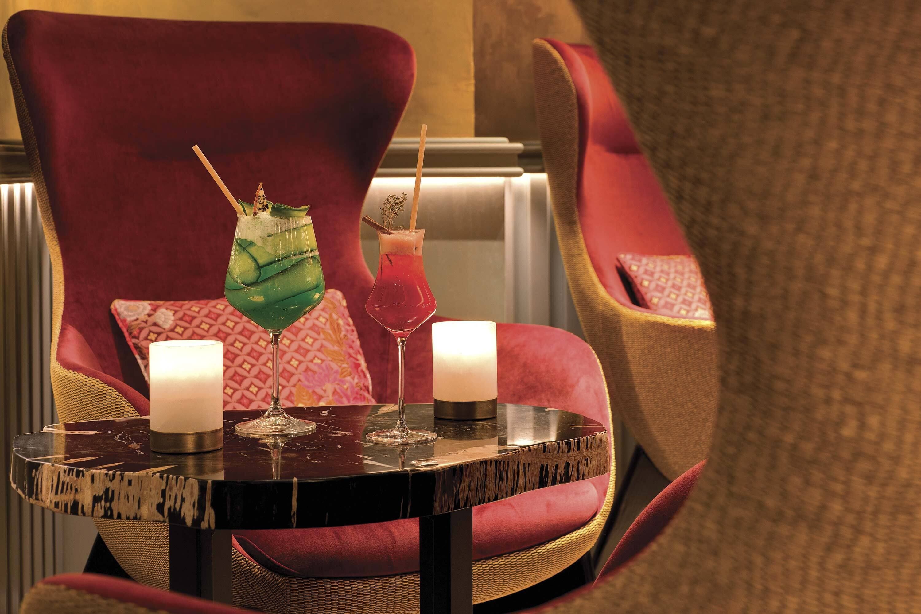 Un fauteuil rouge recouvert de tissu, avec une petite table ronde placée devant, sur laquelle sont posées deux bougies et deux boissons.