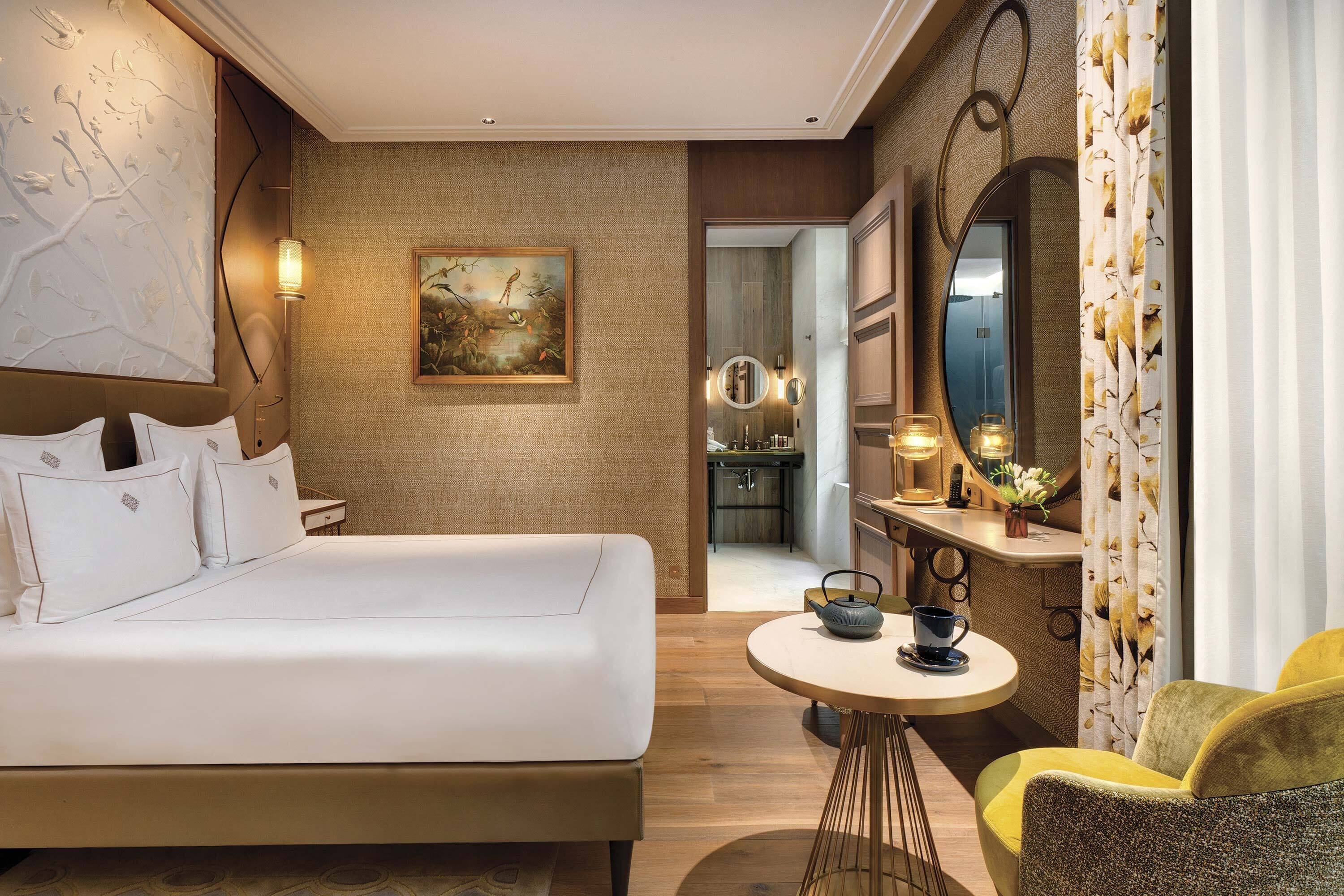 A modern hotel room with a large bed, a small round table with a teapot, a mirror, and a painting on the wall.