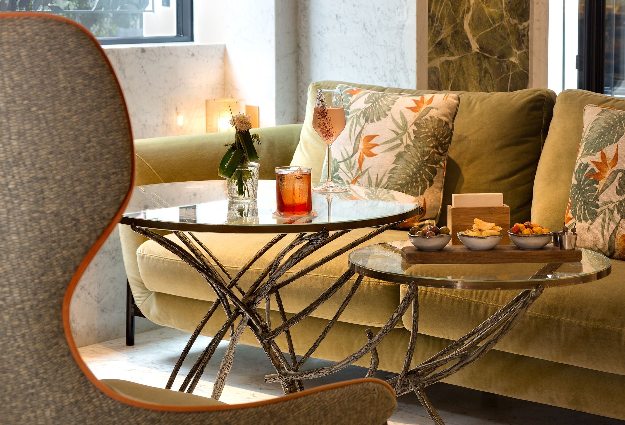 A cozy seating area with a round glass table, a bottle, a glass, and bowls of snacks.