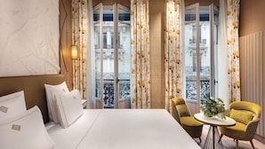 A hotel room with a bed, two chairs, a small table with a vase, and a balcony view. - La Demeure Montaigne (Paris)