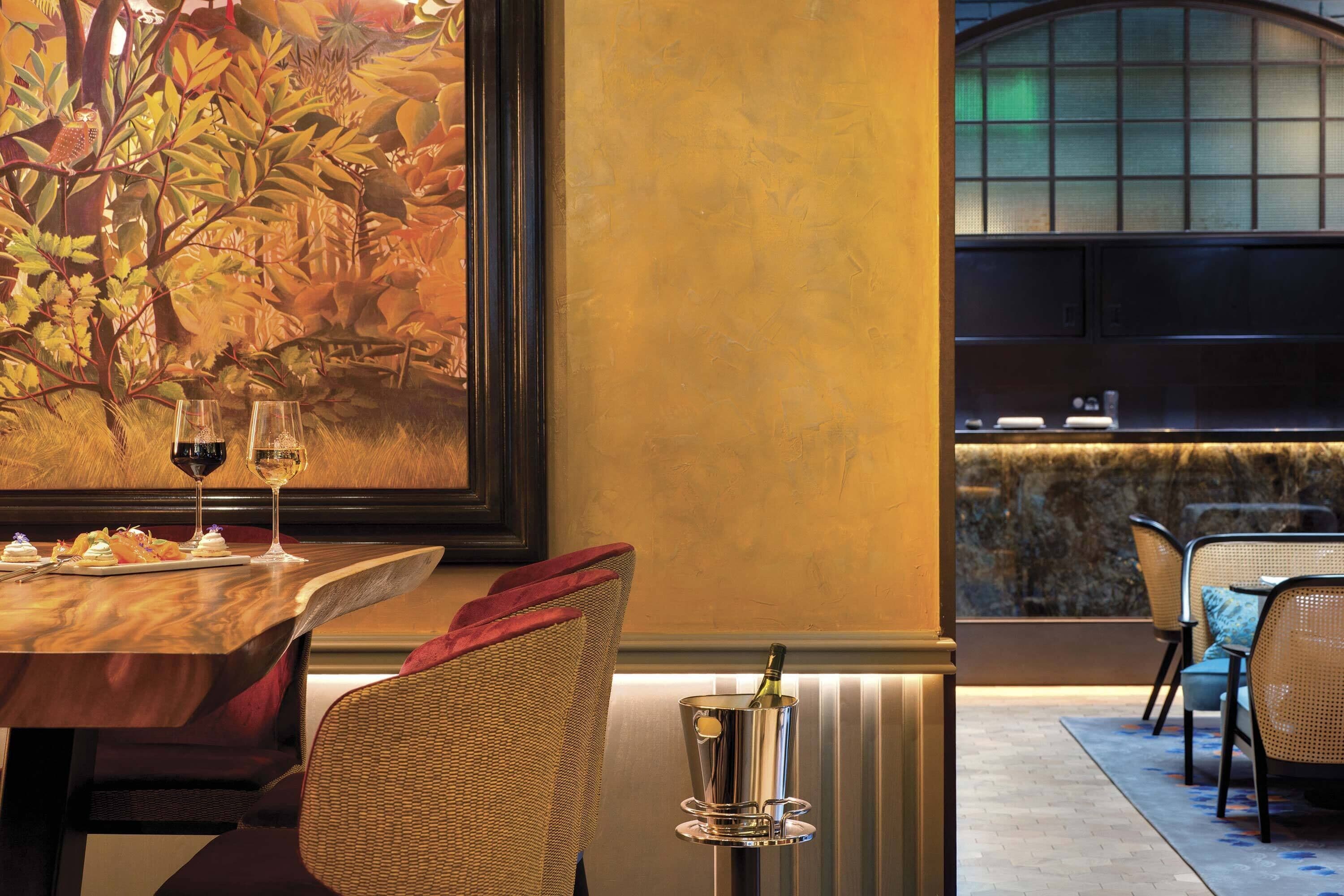 A restaurant with a wooden table, two wine glasses, a bottle of champagne, and a large painting on the wall.