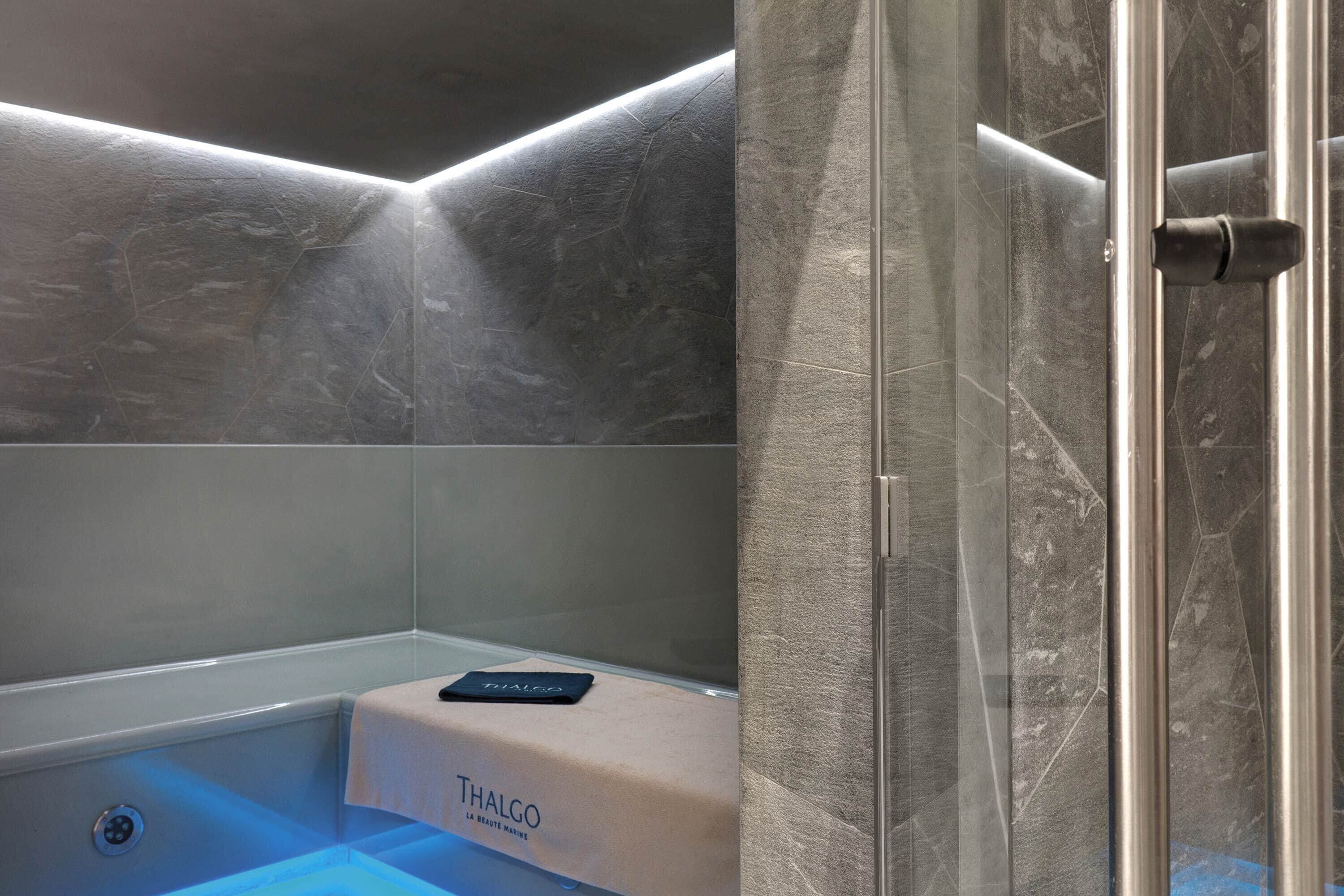 Casa de banho moderna com cabine de duche em vidro, painel de controlo visível e toalheiro da marca Thalgo.