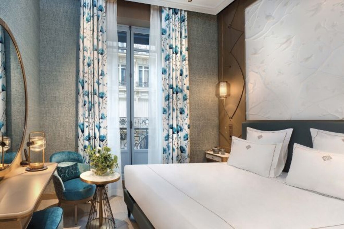 A hotel room with a bed, a desk with a chair, a small table with a plant, and curtains with a floral pattern.