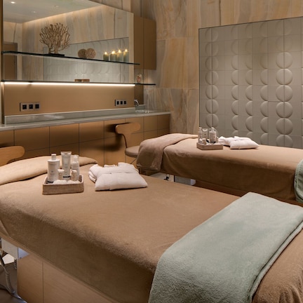A modern spa room with two massage tables, a large mirror, and a shelf with toiletries.