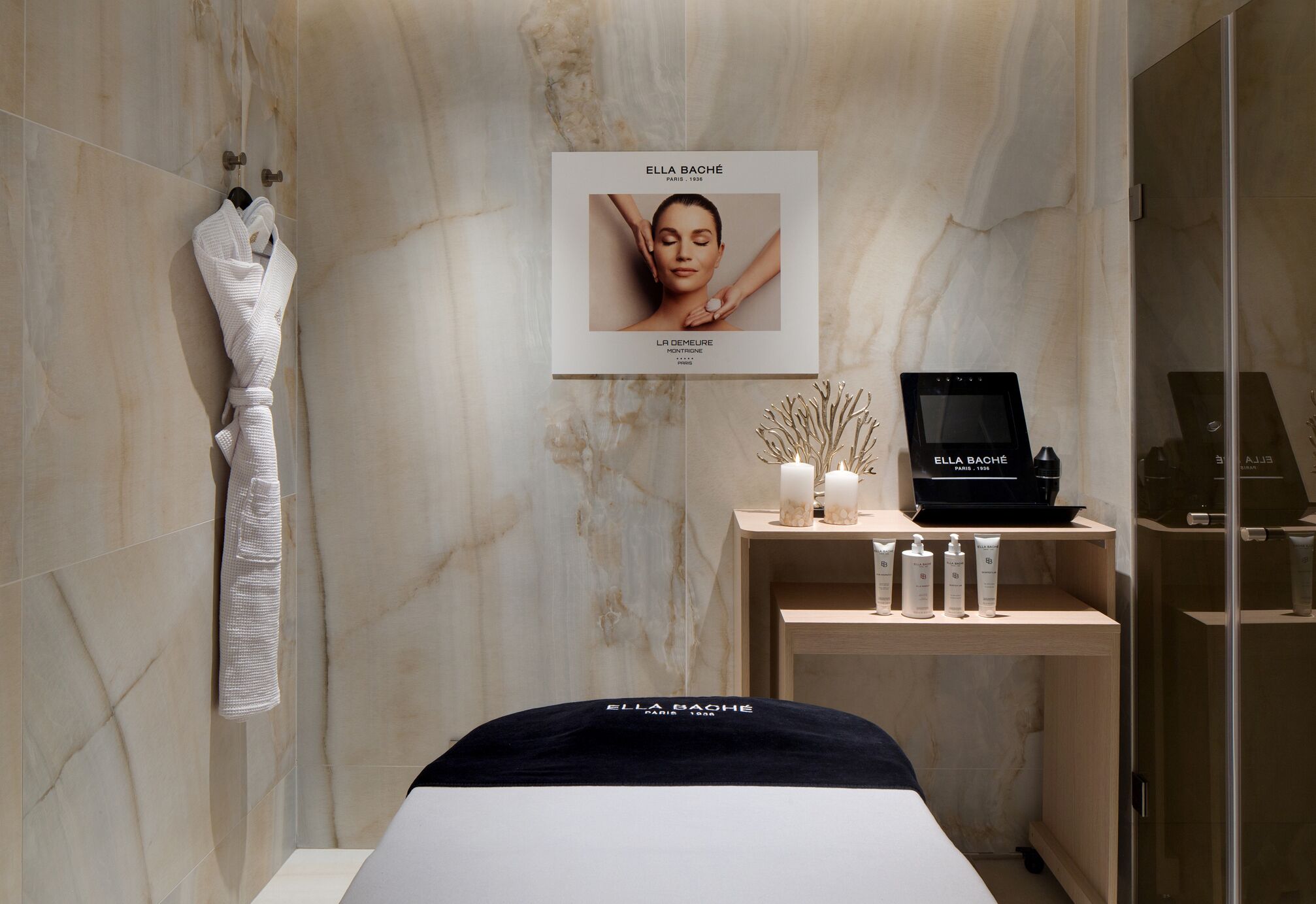 Ein moderner Wellnessraum mit Marmorwand, einem gerahmten Poster, einem Bademantel, einer Holzeinlege mit Kerzen und Hautpflegeprodukten sowie einem schwarzen Massagetisch.