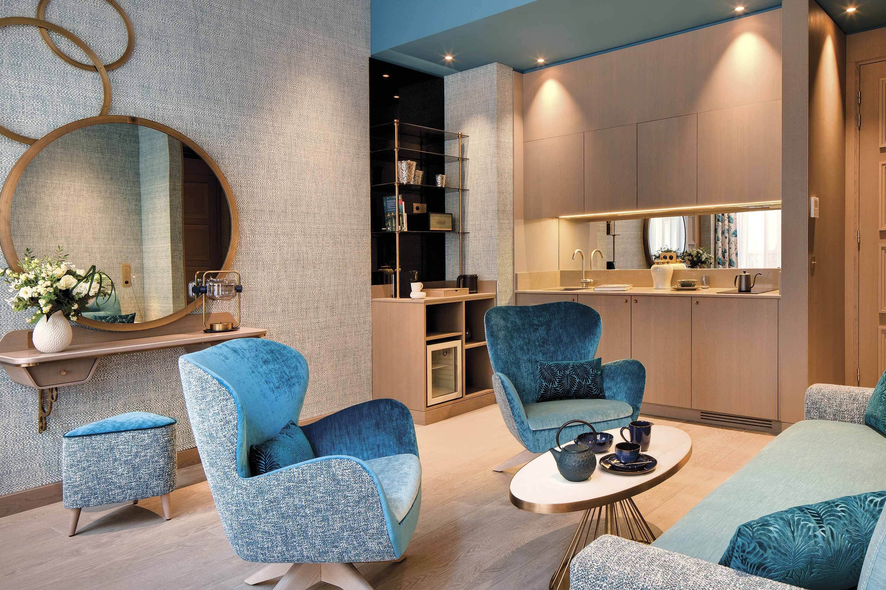 A modern interior with a round mirror, blue upholstered chairs, a small round table, and a kitchen area with a countertop and cabinets.