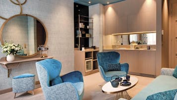 A modern interior with a round mirror, blue upholstered chairs, a small round table, and a kitchen area with a countertop and cabinets.