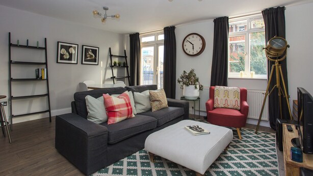 Un salon moderne avec un canapé gris, un fauteuil rouge, un pouf blanc, une bibliothèque, une horloge et un télescope.
