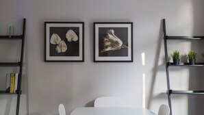 Un espace repas moderne avec une table et des chaises blanches, deux œuvres d’art florales encadrées au mur, et des étagères noires contenant des livres et des plantes.
