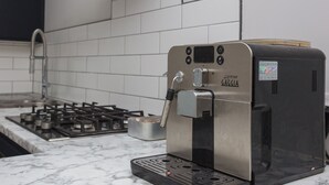 Une cuisine équipée d’une machine à café, d’une cuisinière à gaz et de murs recouverts de carreaux blancs.