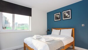 Un lit bien fait, avec du linge de lit blanc et un plaid gris, une fenêtre avec un store noir et deux tableaux encadrés sur le mur bleu.