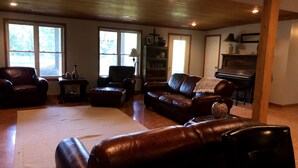 Ein geräumiges Wohnzimmer mit Ledersofas, einem Klavier und großen Fenstern, die natürliches Licht hereinlassen.