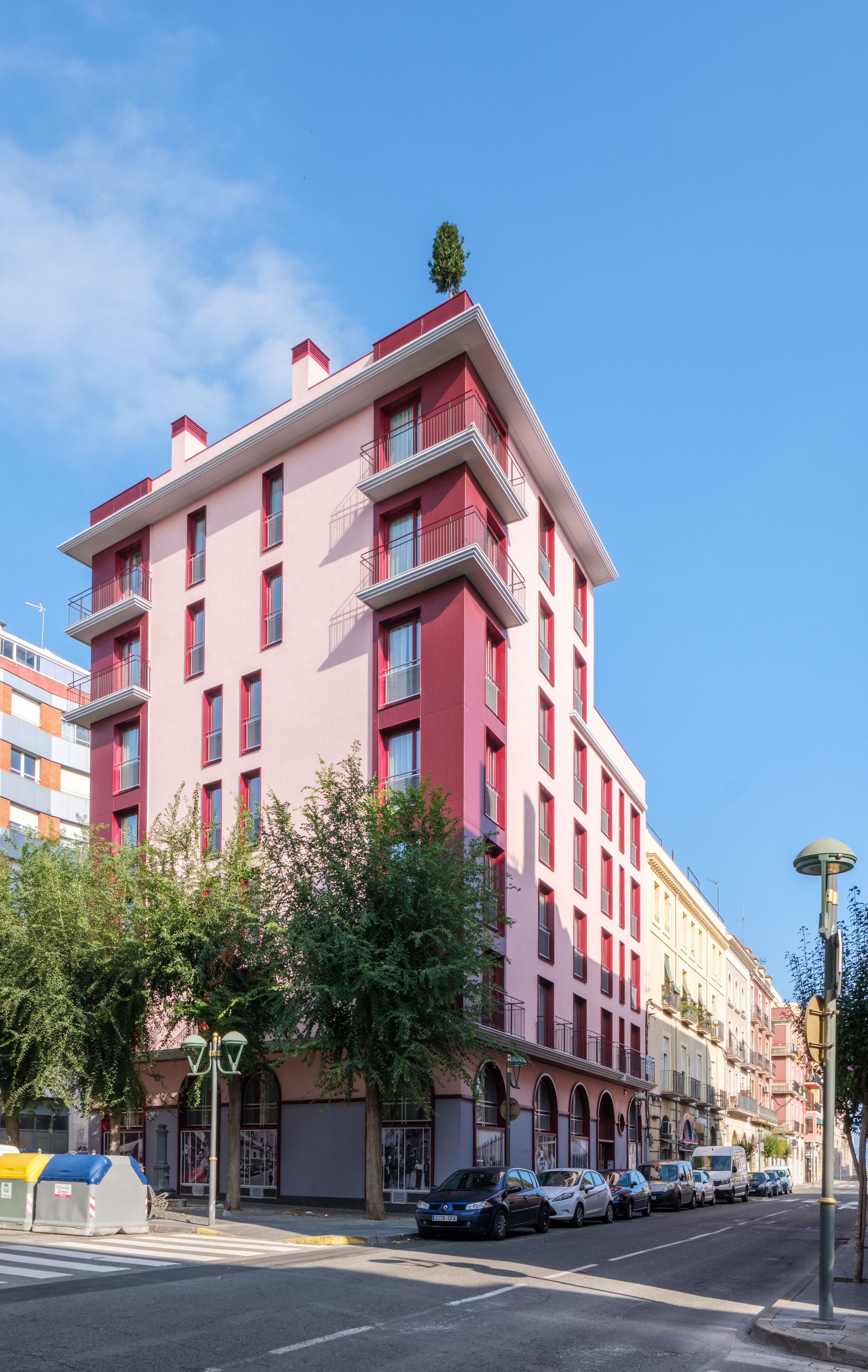 Ett hotell med flera våningar och balkonger, en klarblå himmel och parkerade bilar längs gatan.