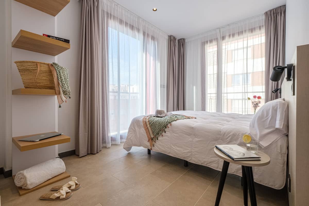 Quarto bem arrumado com cama, mesa de cabeceira, um laptop numa prateleira e janela com cortinas.