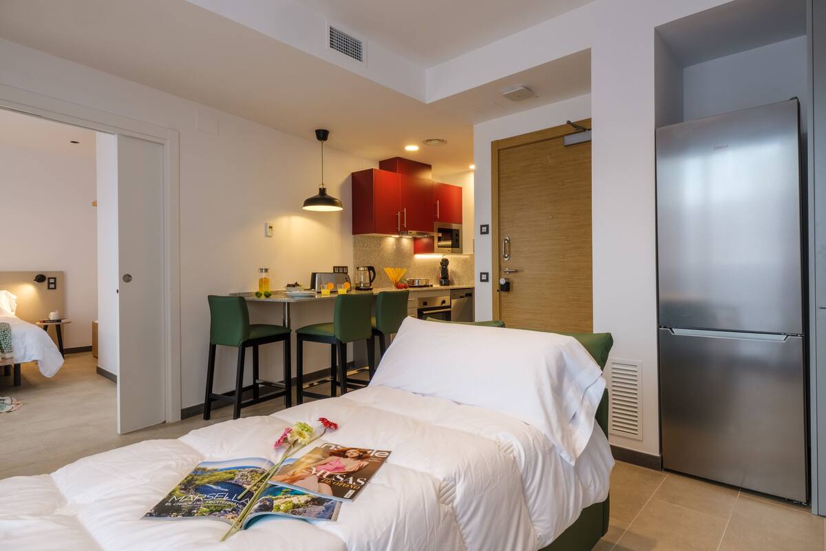 Quarto de hotel compacto com cama, pequeno espaço de cozinha, área de refeições e banheiro separado.