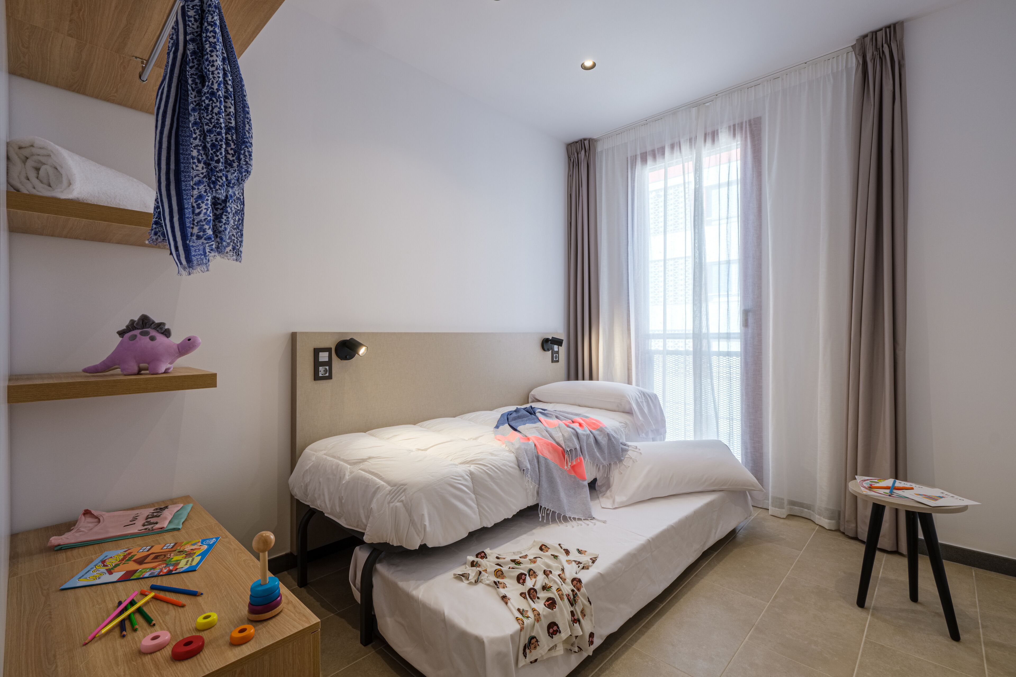 A neatly arranged bedroom with a bed, a small table with children's toys, and a shelf with a stuffed toy.