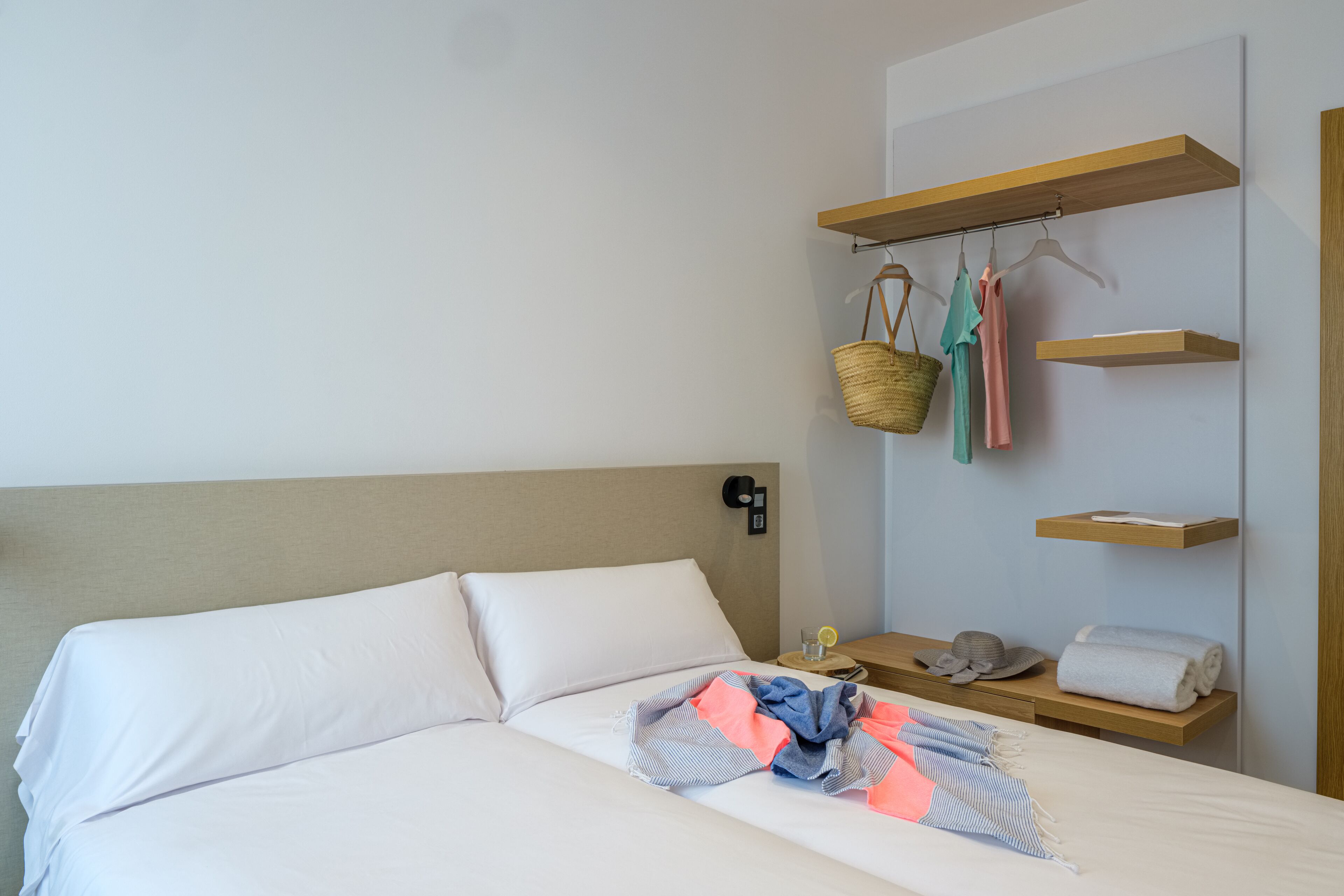 A neatly made bed with white linens, a bedside table with a lamp, a hanging basket, and a shelf with folded towels and a hat.