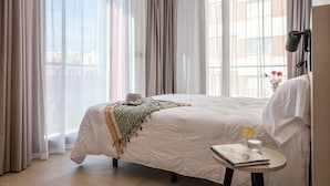 Habitación de hotel con una cama grande, una mesita de noche con un libro y un vaso de agua, y vista a la ciudad a través de cortinas translúcidas.