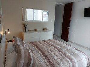 Ein Schlafzimmer mit Bett, Nachttisch, Lampe, Kommode und einem Fenster mit Jalousien.
