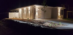Ett modernt hus i mörkret, med en väl upplyst uppfart och ett litet träd på framsidan av tomten.