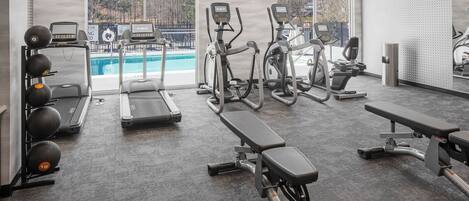 A gym with treadmills, elliptical machines, and weightlifting equipment. There is a pool visible through the windows.