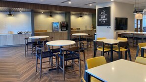A modern cafe interior with round tables and chairs, a counter with a beverage dispenser, and a wall-mounted TV.