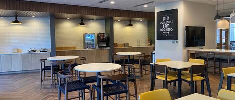 Interior de una cafetería moderna con mesas y sillas redondas, una barra con dispensador de bebidas y un televisor montado en la pared.