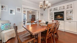 A dining area with a wooden table and chairs, a chandelier, and a fireplace with shelves above.