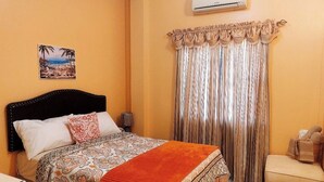 A bedroom with a bed, a nightstand, a chair, a wall-mounted air conditioner, and a curtain. - Cozy 2 Bedroom Apt near Airport, Malls, Golfing, Mountains  (Arouca)