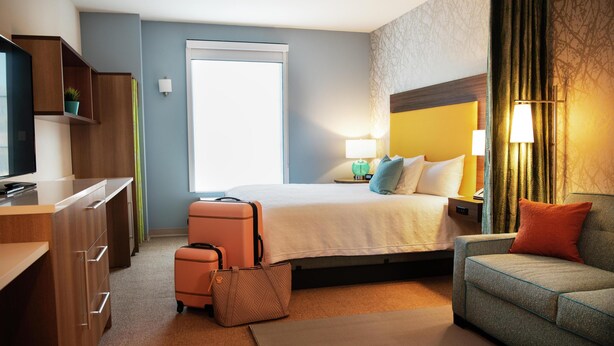 A hotel room with a bed, a sofa, a desk, and a suitcase. - Home2 Suites by Hilton Hayward (Hayward)
