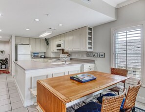 A modern kitchen with white cabinetry, a wooden dining table, wicker chairs, and a sign indicating a beach direction.