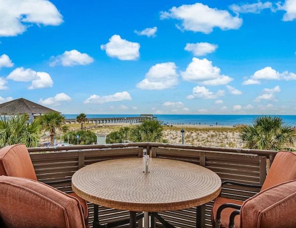 A round table with a mosaic top, surrounded by cushioned chairs, overlooking a beach with a pier in the distance.