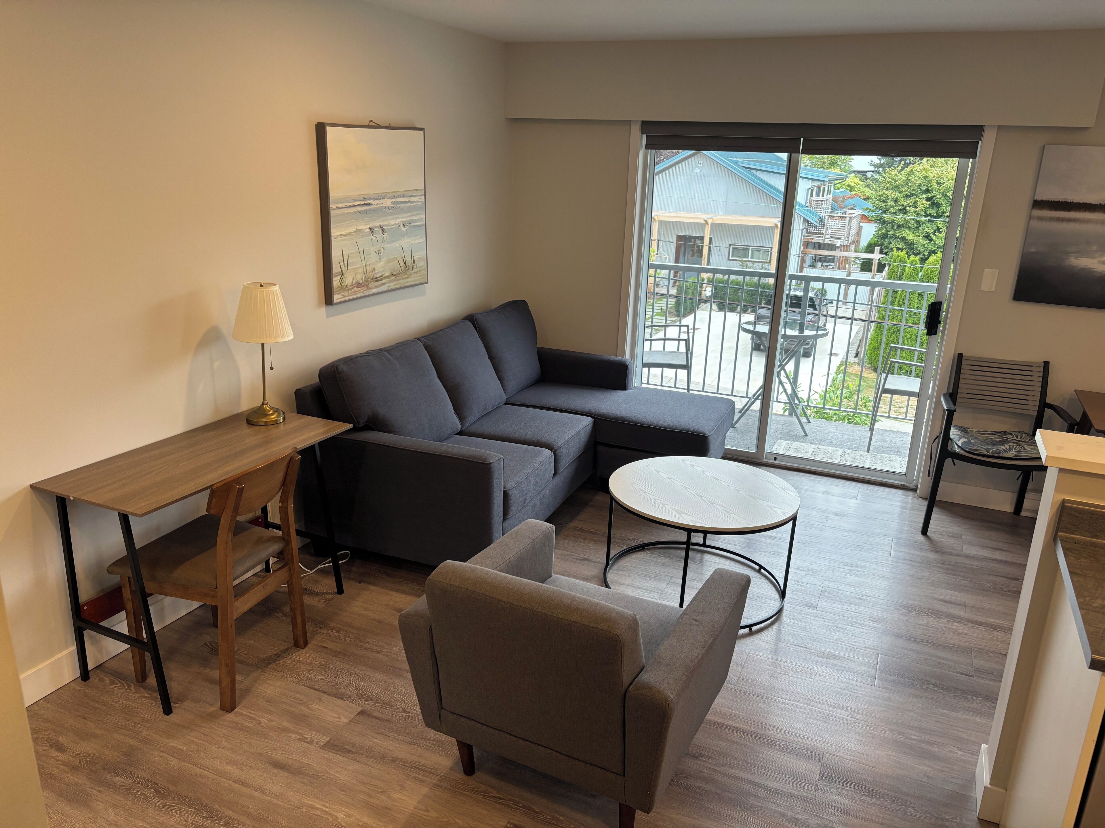 A modern living room with a sofa, a round coffee table, a desk, and a chair. There is a large window with a view of the outside.