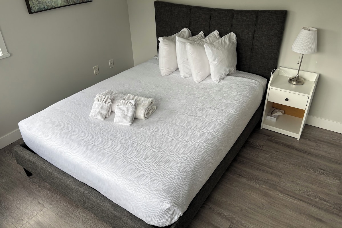 A neatly made bed with white linens and pillows, a bedside table with a lamp, and a small drawer unit.