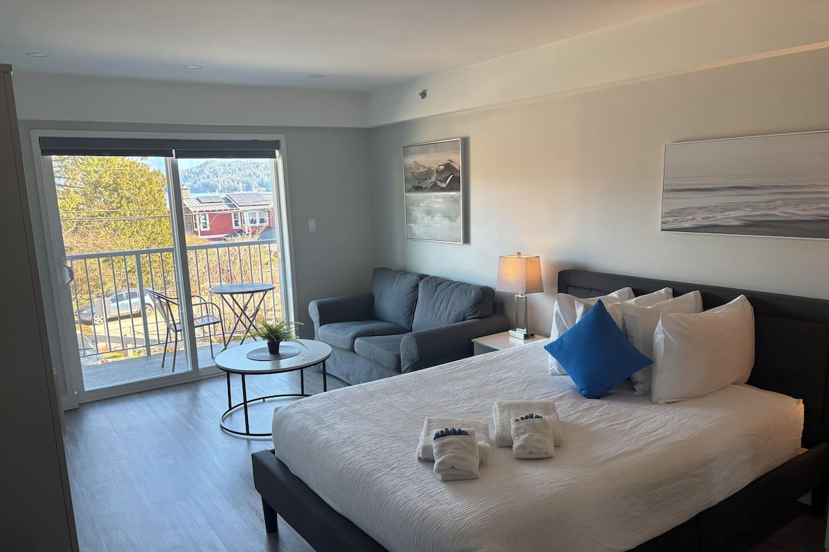 A hotel room with a bed, a sofa, a small table, a lamp, and a view of the outside.