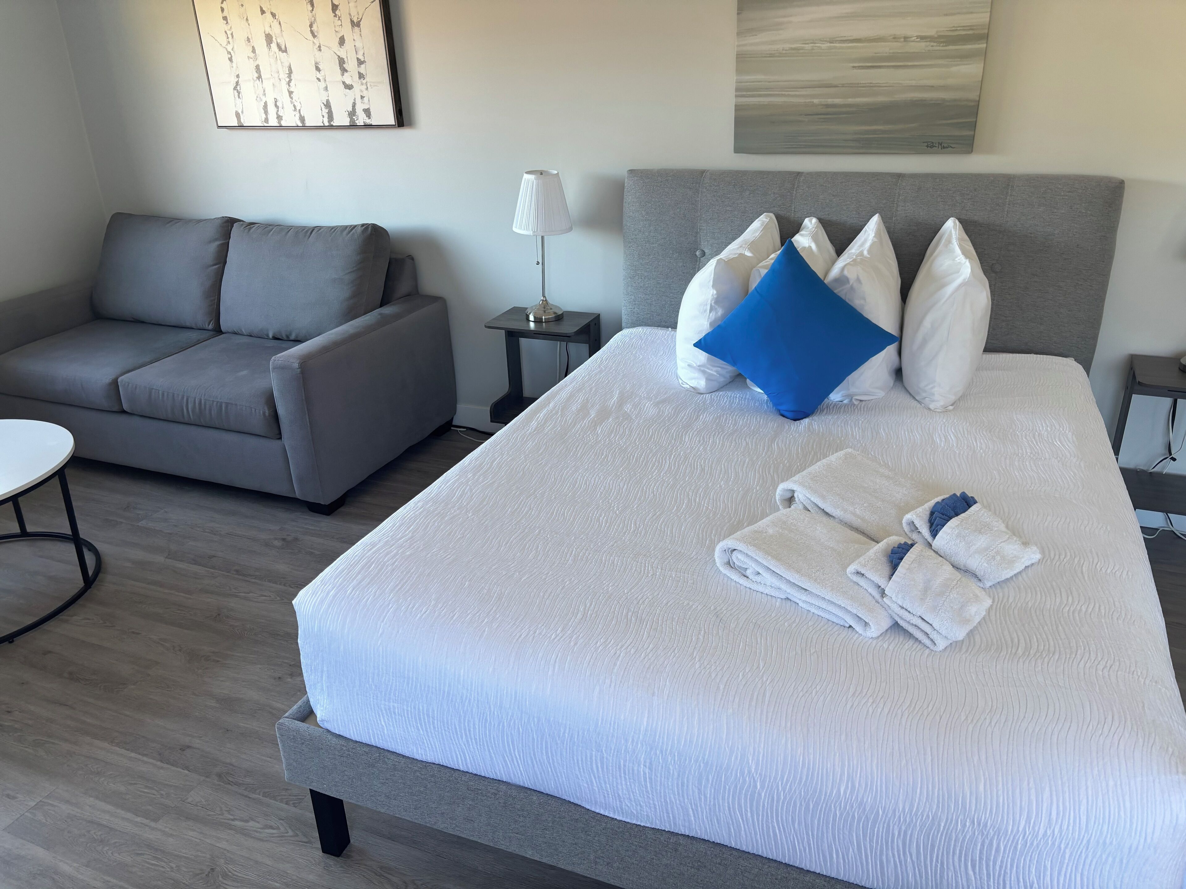 A neatly made bed with white linens and a blue pillow, a grey sofa, and a small table with a lamp.