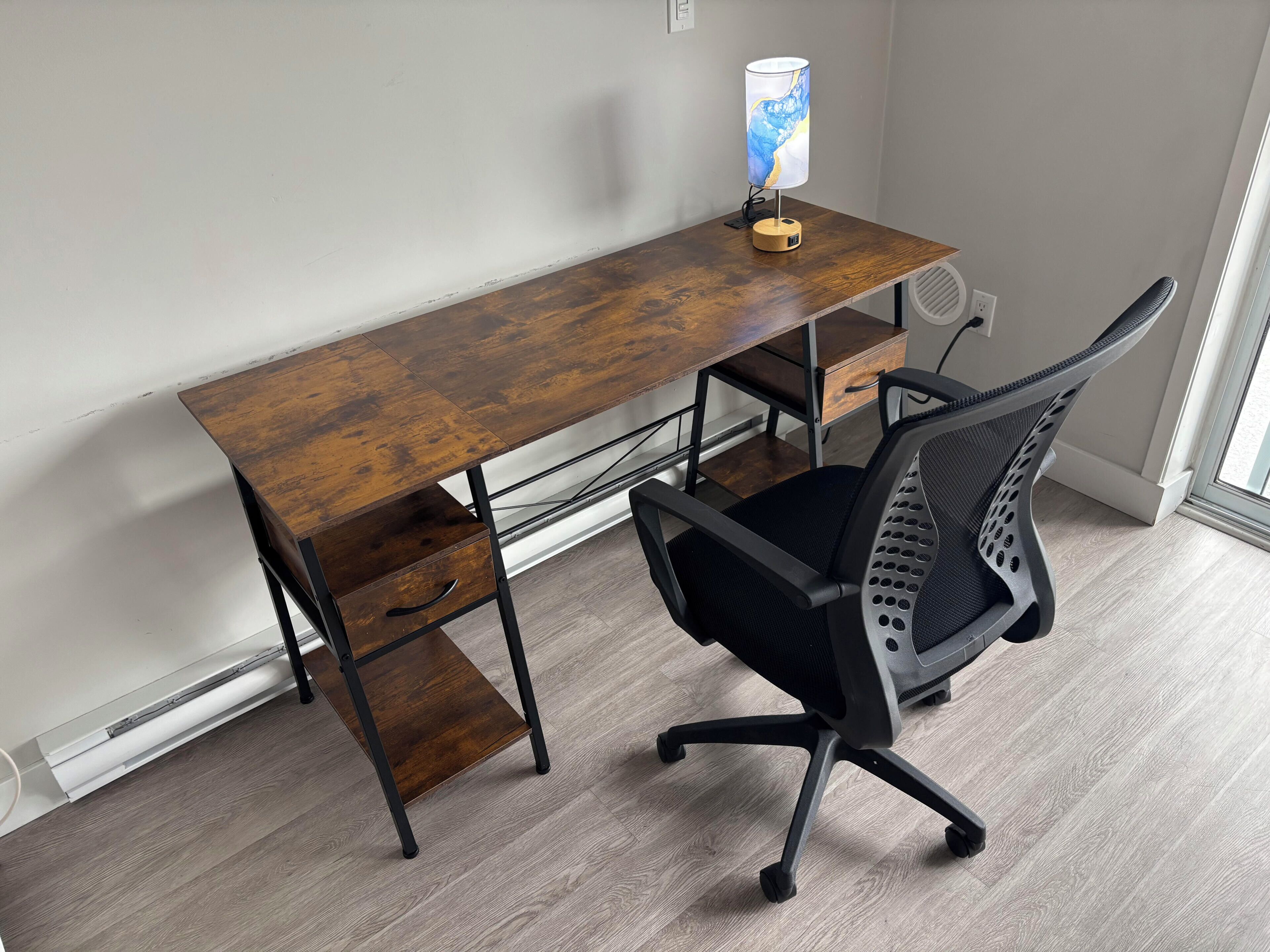 A wooden desk with a lamp, a black office chair, and a window with blinds.