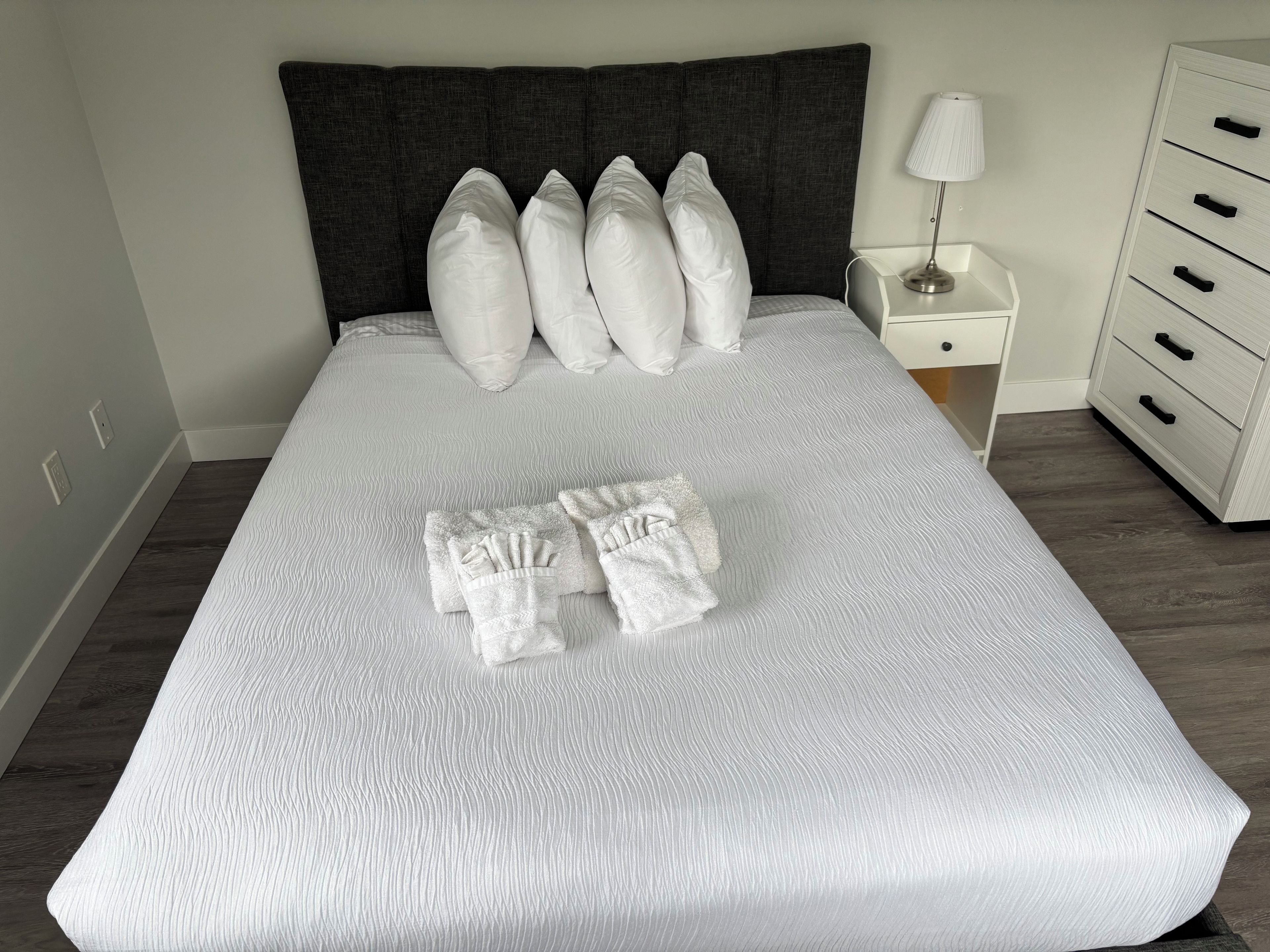 A neatly made bed with white bedding and pillows, a bedside table with a lamp, and a white dresser with drawers.