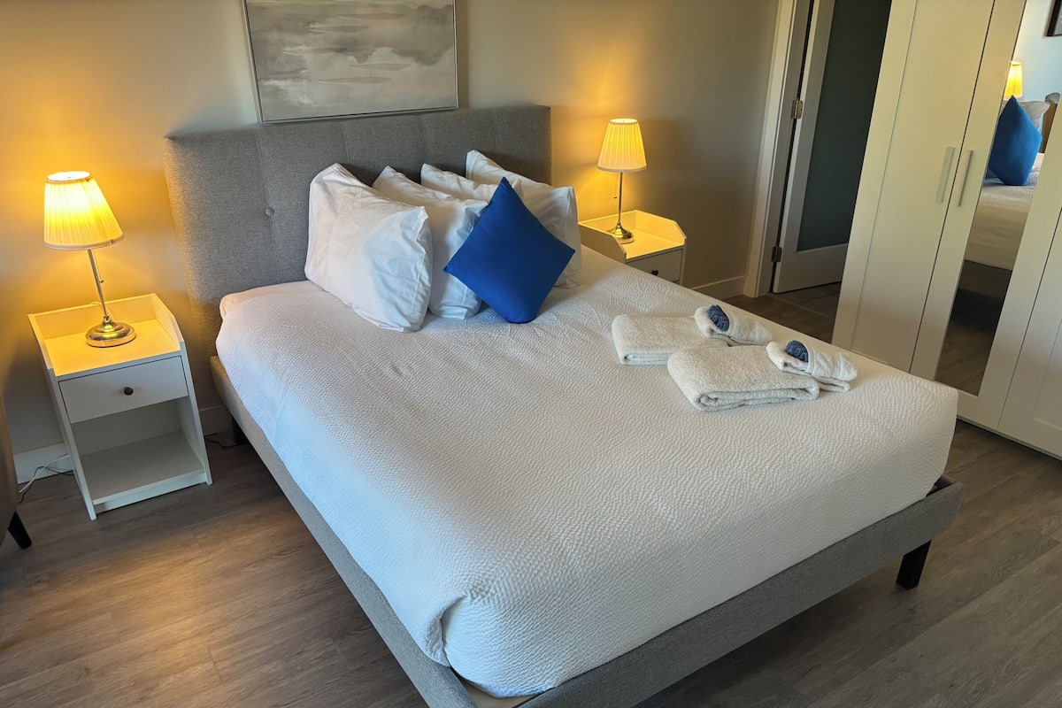 A neatly made bed with pillows, two bedside lamps, and folded towels on a wooden floor.