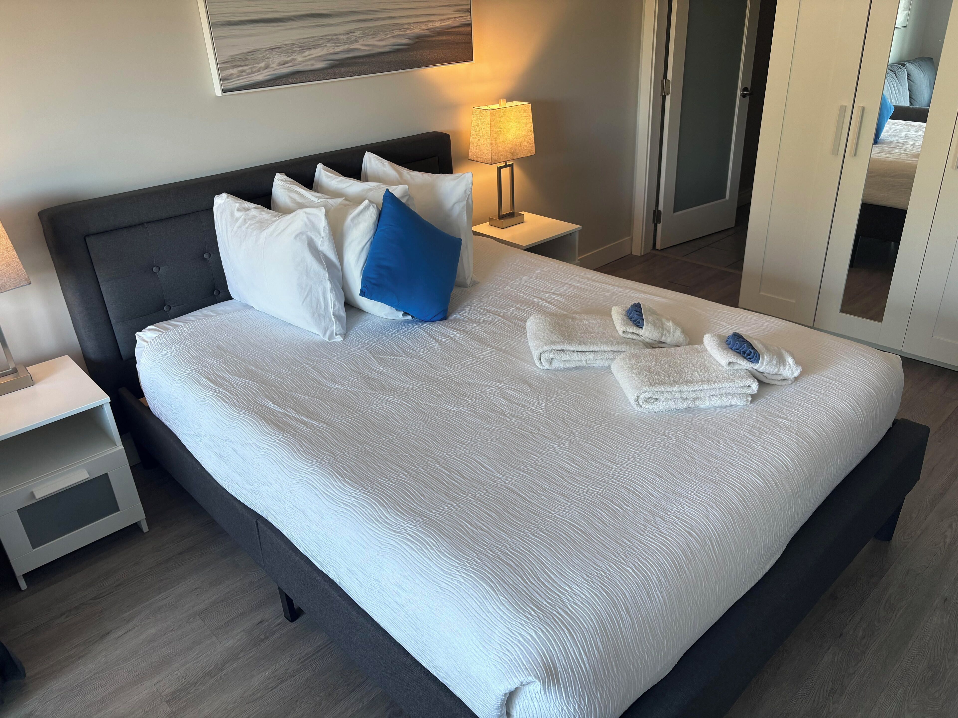 A neatly made bed with white and blue pillows, a white textured bedspread, and two folded towels on the bed.