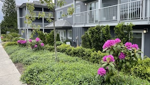 Un edificio residencial de varios pisos con balcones, un jard ín bien cuidado con flores rosadas y un cielo despejado.