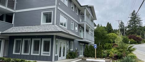 A multi-story building with a grey facade, multiple balconies, and a parking area with a handicapped parking sign.