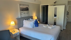 Una cama bien hecha, con almohadas, una lámpara de noche y una mesita de noche. Se observa un armario con espejo y una puerta que da a un baño.