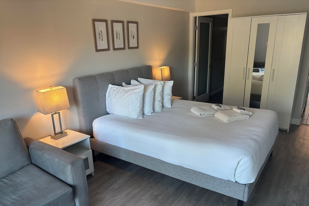 A modern hotel room with a large bed, a bedside table with a lamp, a grey armchair, and a wardrobe.