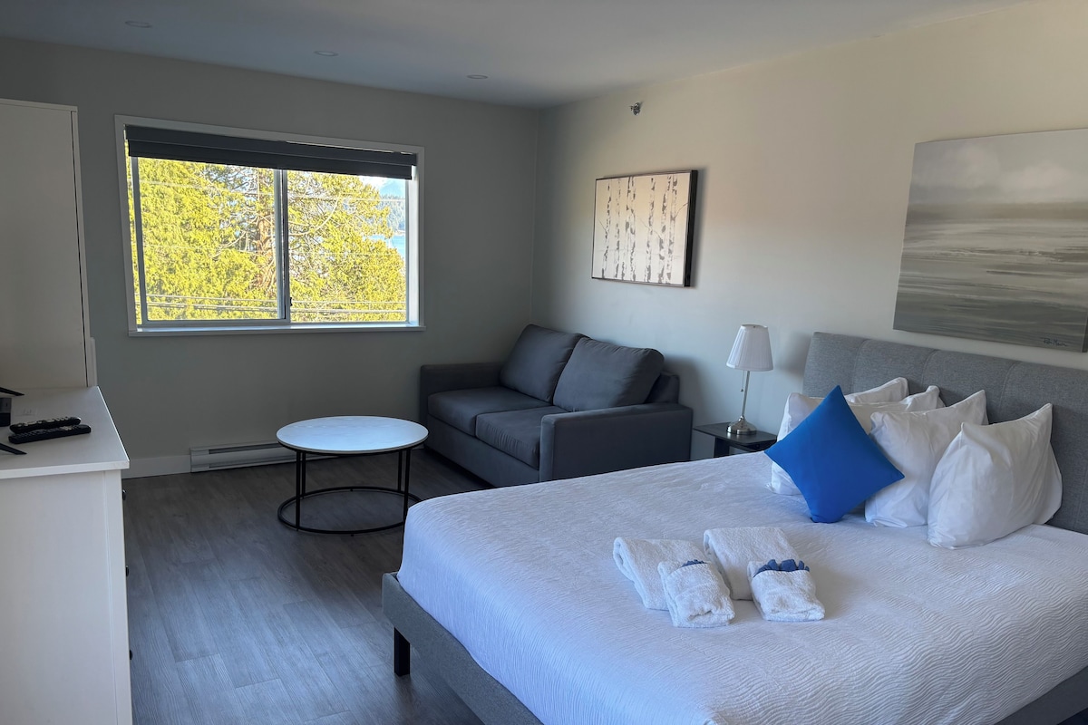 A modern hotel room with a large bed, a grey sofa, a small round table, and a window with a view of trees.